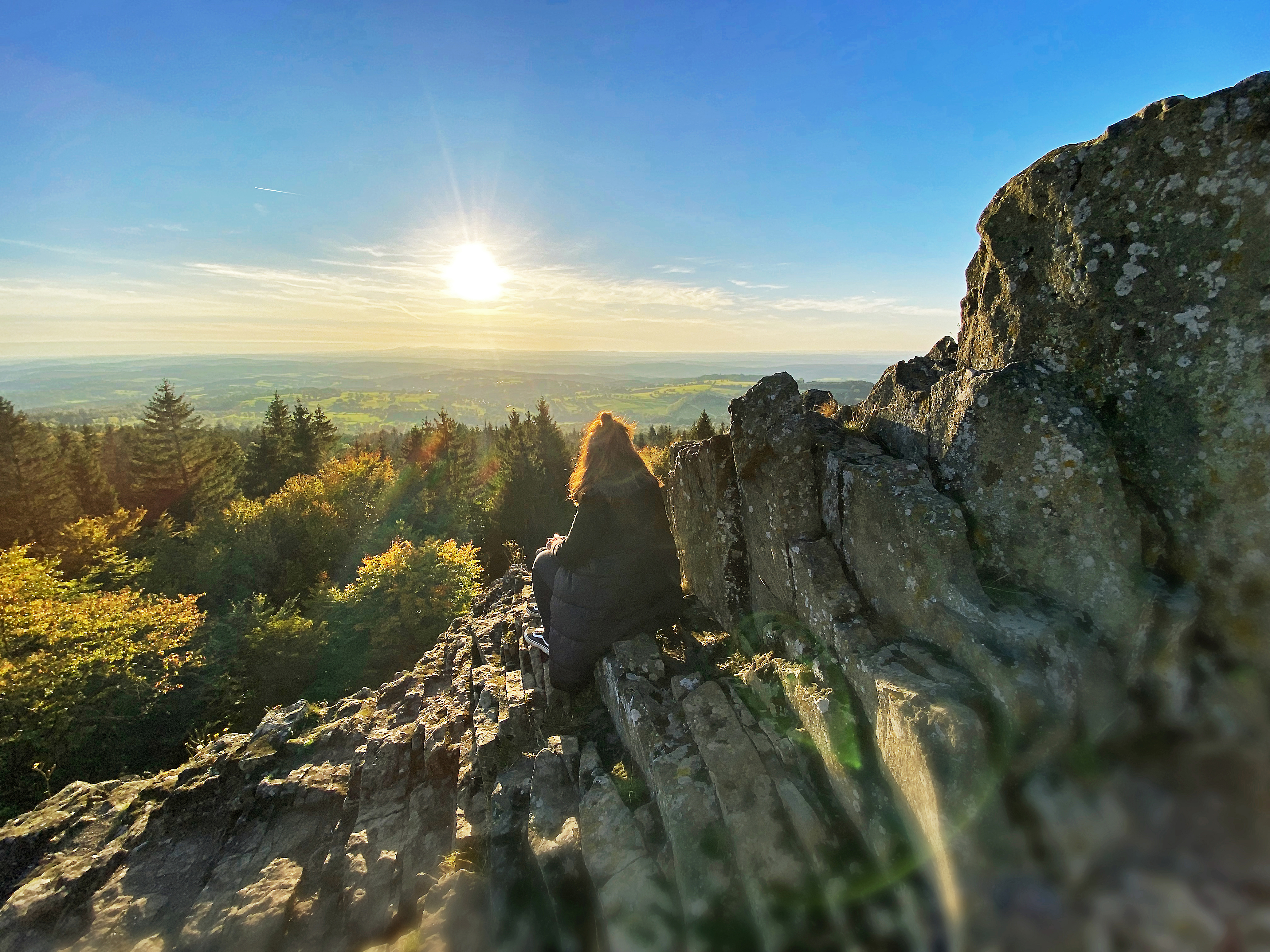 Der Blick des verweilenden Wanderers auf dem schroffen Basaltfelsen des Geotopes Bilstein im hessischen Vogelsberg geht weit über das geschwungene Land der Vulkanregion. Der Herbst hat das Laub des Waldes bunt gefärbt und die warmen Sonnestrahlen zaubern ein magisches Licht am strahlend blauen Himmel.